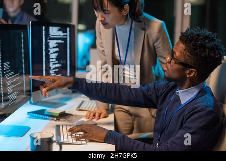 Two young intercultural colleagues discussing coded data on computer screen while African businessman pointing at it Stock Photo