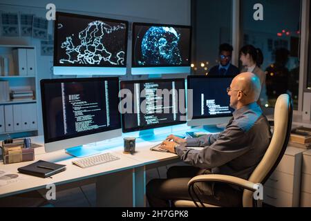 Bald businessman in formalwear sitting in front of computer monitor and decoding data with two colleagues on background Stock Photo