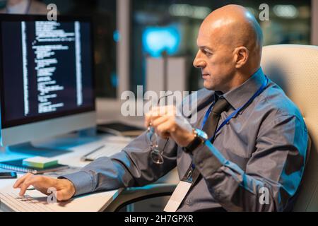 Mature bald it-engineer working in front of computer monitor while sitting in armchair by desk in openspace office Stock Photo