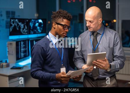 Two elegant intercultural businessmen discussing data in digital tablet held by mature programmer or it-engineer Stock Photo