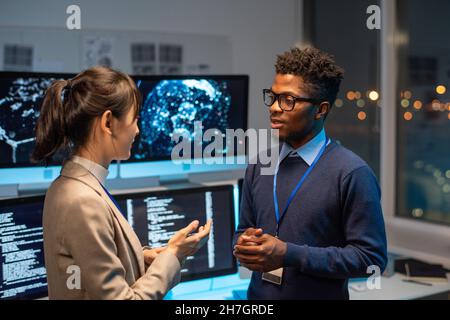 Two young multiracial colleagues having discussion against workplace with computer monitors in large openspace office Stock Photo