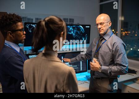 Bald mature director of software company giving instructions to young intercultural subordinates while pointing at data on computer screen Stock Photo