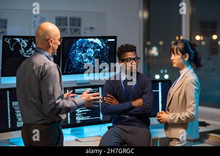 Three intercultural colleagues in formalwear discussing working points at meeting by their workplace in openspace office Stock Photo