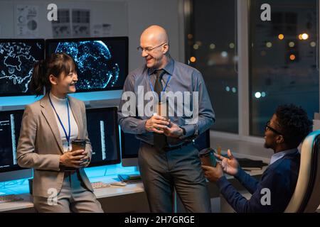 Three happy colleagues with drinks having discussion while sitting by desk with computer monitors in office environment Stock Photo