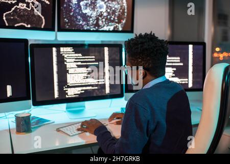 Young African male software developer working late in front of computer screen with coded data in office environment Stock Photo