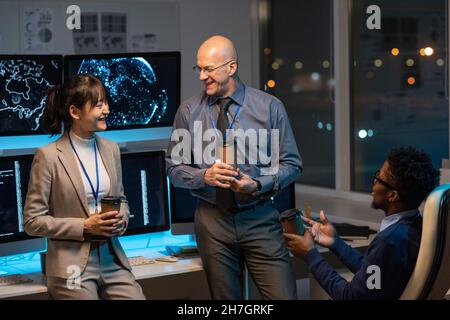 Three happy colleagues with drinks having discussion while sitting by desk with computer monitors in office environment Stock Photo