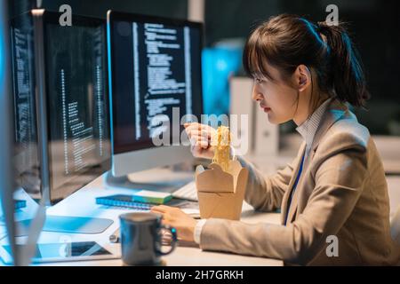 Side view of young Asian female programmer eating chinese wok during work over coded data in front of computer monitor Stock Photo