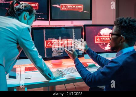 Young African male programmer complaining female colleague about problem in work of computers while sitting by workplace Stock Photo