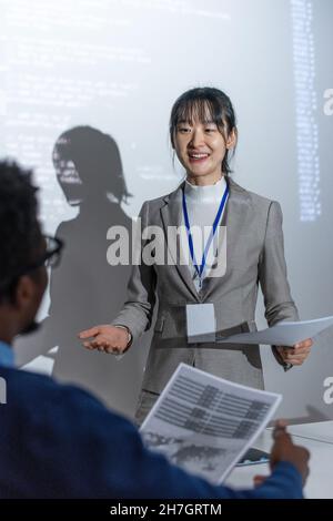 Happy young Asian female with papers standing by interactive whiteboard with data while speaking to African male colleague at seminar Stock Photo