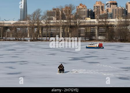 Saint-Petersburg, Russia - March 24, 2021: Man is engaged in winter fishing on the ice of the river and a hovercraft Emercom passes by against the bac