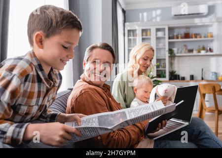 boy reading newspaper near dad working with laptop and mom playing with brother on sofa Stock Photo