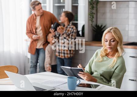 woman writing in notebook while working in kitchen near husband and sons on blurred background Stock Photo