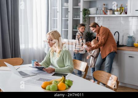 busy woman writing in notebook while working near laptop and blurred family in kitchen Stock Photo