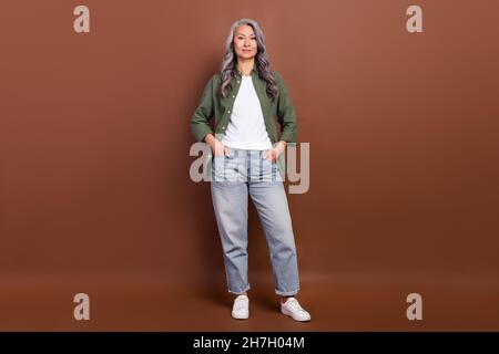 Full size photo of cool old white hairdo lady stand wear khaki shirt jeans sneakers isolated on brown color background Stock Photo