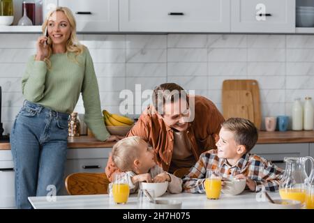 cheerful man talking to sons having breakfast near wife with mobile phone on blurred background Stock Photo