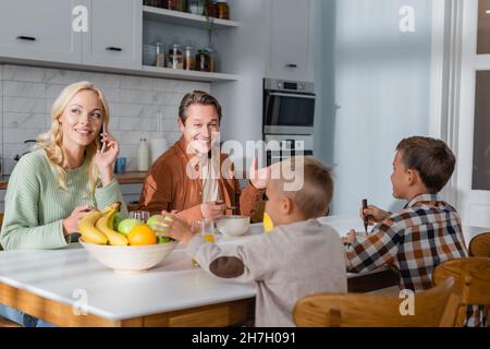 cheerful man talking to sons during breakfast while wife calling on mobile phone Stock Photo