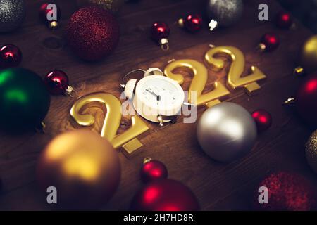 Candles in the form of numbers 2022, alarm clock showing midnight, multicolored christmas balls on a wooden background. Dark Christmas background. Stock Photo