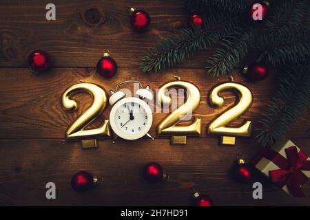Candles in the form of numbers 2022, alarm clock showing midnight, branches, christmas balls and a gift on a wooden background. Dark Christmas backgro Stock Photo