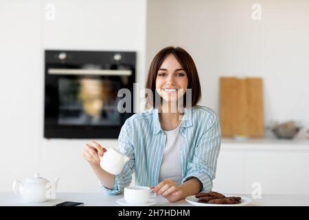 Morning breakfast. Happy woman enjoying coffee and pouring milk in cup, sitting in kitchen and smiling at camera Stock Photo