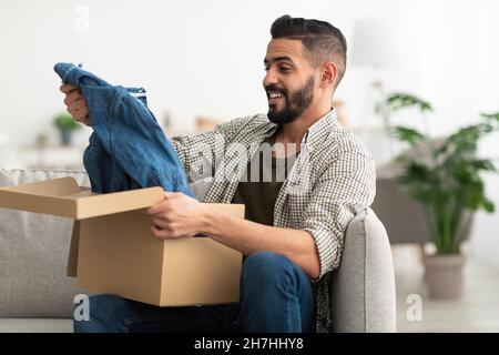 Happy Arab man taking out new clothes from carton box, satisfied with delivery service at home Stock Photo