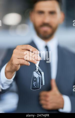 Blur background of caucasian male customer standing at car salon, holding keys from new car and showing thumb up. Focus on male hands with keys.  Stock Photo