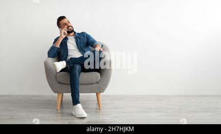 Happy young Arab man talking on smartphone, sitting in armchair against white studio wall, free space Stock Photo