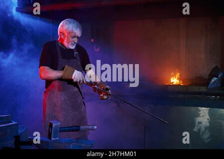 Skillful caucasian blacksmith in protective apron and gloves making sharp sword at forge. Industrial work in workshop of blacksmith. Manual forging concept. Stock Photo
