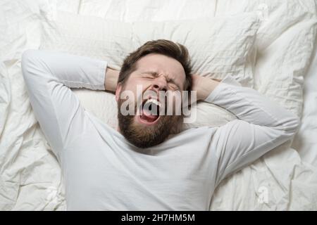 Cute bearded man wakes up from sleep, he yawns and stretches as he lies in bed. Top view. Close-up. Stock Photo