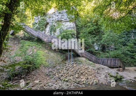 Staircase with wooden fence between sandstone rocky mountain wall and the Black Ernz river, abundant wild vegetation in the background, sunny summer d Stock Photo