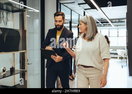 Colleagues having an important discussion while walking together in an office. Two diverse businesspeople using a digital tablet in a modern workspace Stock Photo