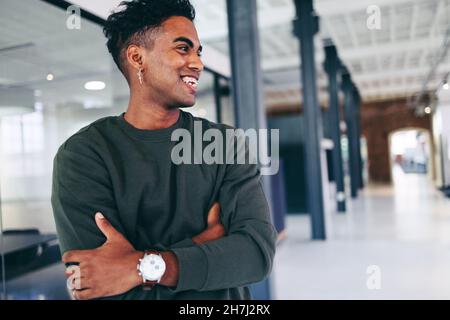 Modern businessman smiling while standing alone in a creative office. Happy young businessman looking away cheerfully while standing with his arms cro Stock Photo
