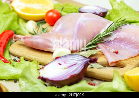Raw uncooked pink rabbit leg meat with green rosemary and fresh vegetables on light wooden cutting board background. Stock Photo