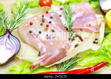 Raw uncooked pink rabbit leg meat with green rosemary and fresh vegetables on light wooden cutting board background. Stock Photo