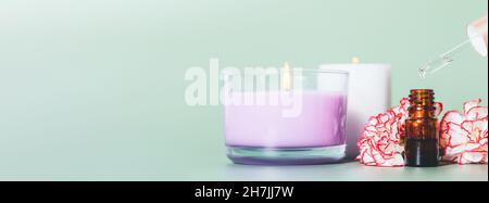 Relax aromatherapy with natural essential oil dripping in bottle next to fresh flowers and burning aroma candles on green background with copy space. Stock Photo