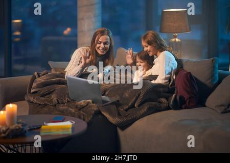 Pretty caucasian mother and her little daughters sitting on couch, smiling and waving hands during video call on modern laptop. Concept of family, technology and online communication. Stock Photo