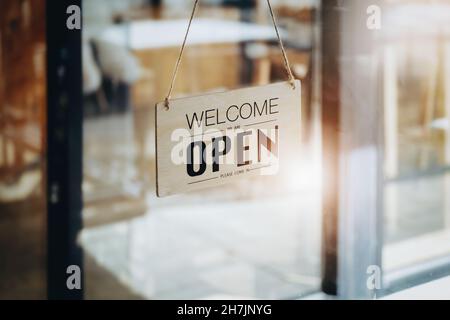 WELCOME WE ARE OPEN PLEASE COME IN notice sign wood board label hanging through glass door. Stock Photo