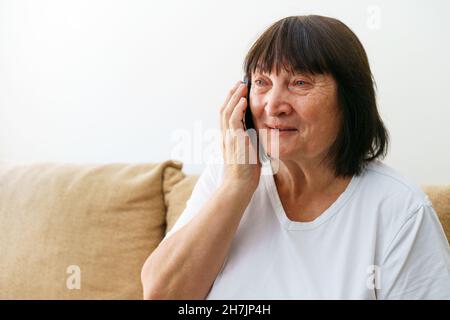 Cellular. Portrait of cheerful caucasian grandmother calling with family while sitting on sofa in modern living room and looking to side. Senior woman talking on the phone having pleasant conversation Stock Photo