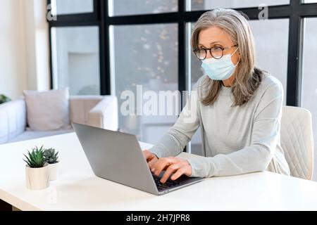 Gray-haired mature businesswoman wearing protective medical mask using laptop sitting at the desk in bright office, senior female employee protects herself against viral disease during pandemic Stock Photo