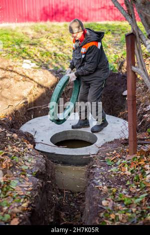 A worker installs a sewer manhole on a septic tank made of concrete rings. Construction of sewage disposal systems for private houses. Stock Photo