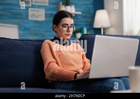 Student influencer on couch holding laptop and typing. Programmer with glasses on sofa writting code in home living room office. Content creator looking at screen posting on social media. Stock Photo