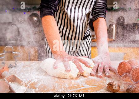 Сloud of white flour flying into air as a woman chef energetically kneads dough on the kitchen table. Homemade non-professional cooking concept. Stock Photo