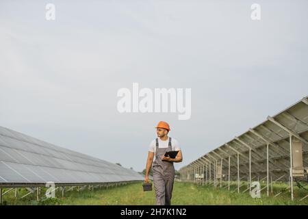 Indian man in uniform and helmet examining work of solar panels while walking on station. Competent engineer carrying clipboard and instruments in hands. Stock Photo