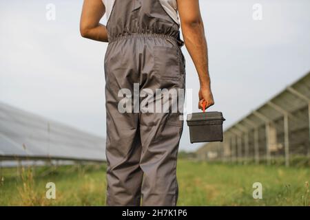 Close up of male engineer in grey overalls with tools in hands standing among solar farm. Man maintaining continuous operation of photovoltaic cells. Green energy concept. Stock Photo
