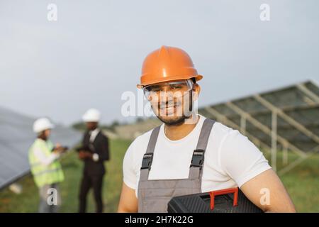 Smiling indian foreman in orange helmet and safety glasses looking at camera with two colleagues talking on blur background. Multiracial people work on with green energy on solar station. Stock Photo