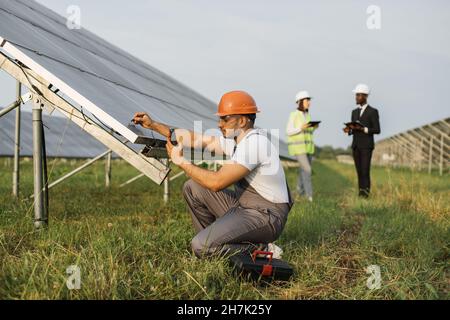 Energy technician in helmet and uniform using multimeter for measuring voltage of solar panels outdoors. Group of engineers with tablet and clipboard standing on background. Stock Photo