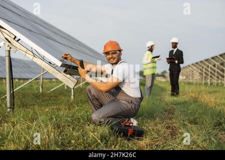 Indian man in uniform squatting near solar panel with multimeter in hands and smiling on camera. Two business people with gadgets talking on background. Stock Photo
