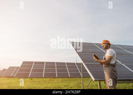 Side view of engineer in orange helmet and brown overalls checking resistance in solar panels outdoors. Indian man using multimeter while working on station. Stock Photo
