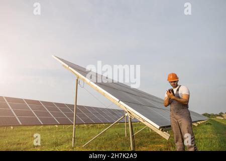 Indian worker in uniform, safety helmet and glasses using multimeter while examining work of solar panels. Concept of people, maintenance and green energy. Stock Photo