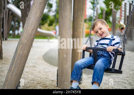 Smiling boy riding on swing at amusement forest park or childish playground having positive emotion Stock Photo