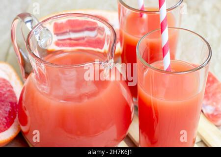 Fresh red grapefruit juice in a glass with a straw and jar with fruit pieces on light wooden background. Stock Photo
